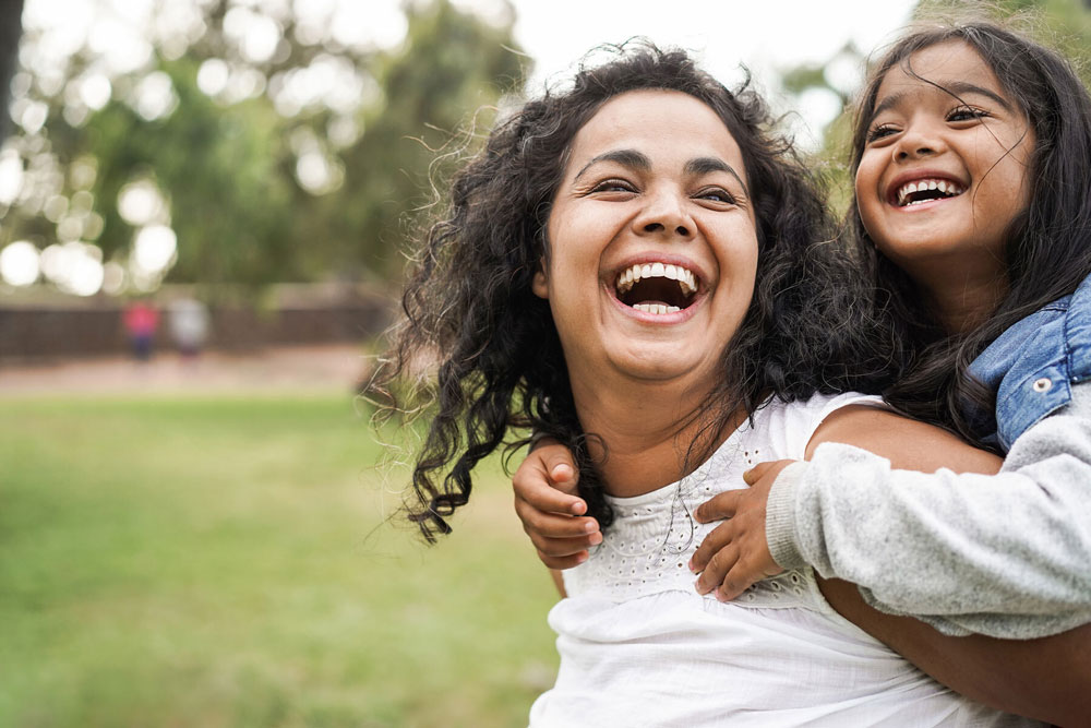 woman smiling with little girl on her back outside in park